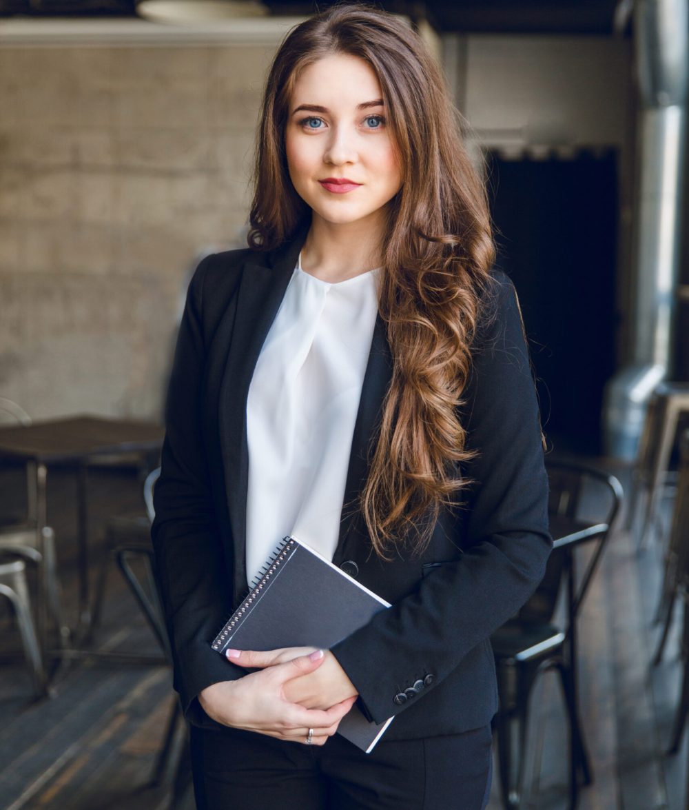 Brunette business woman with wavy long hair and blue eyes stands holding a notebook in hands. She wears a black suit and a white blouse. She is in a cafe with grey walls and dark furniture.